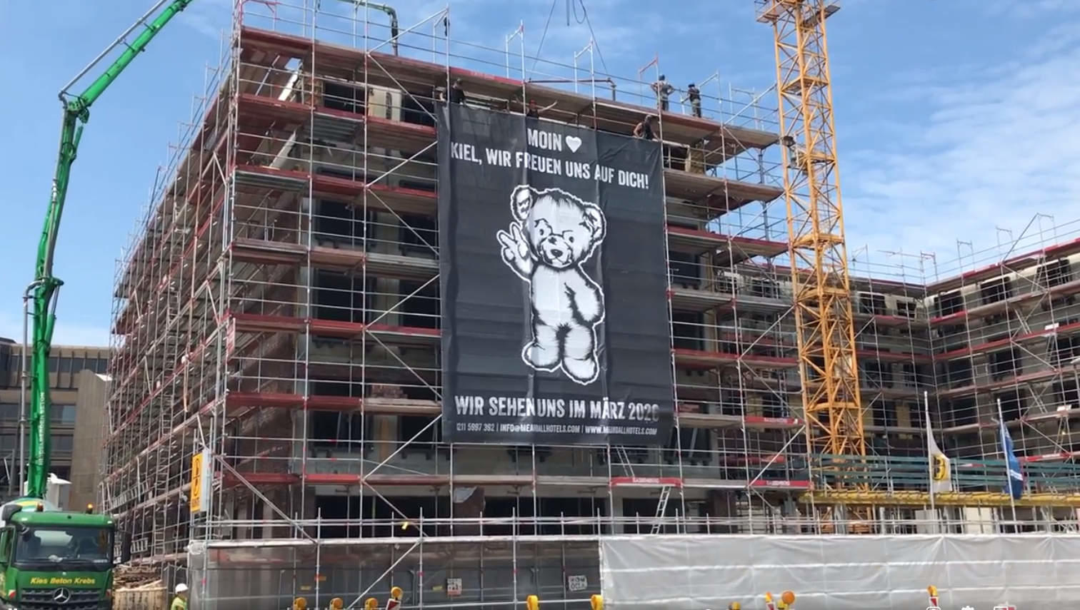 Foto: Baustelle des me and all hotels Kiel mit großem Transparent, das einen stehenden Teddy zeigt, der mit einer 'Victory'-Pose grüßt